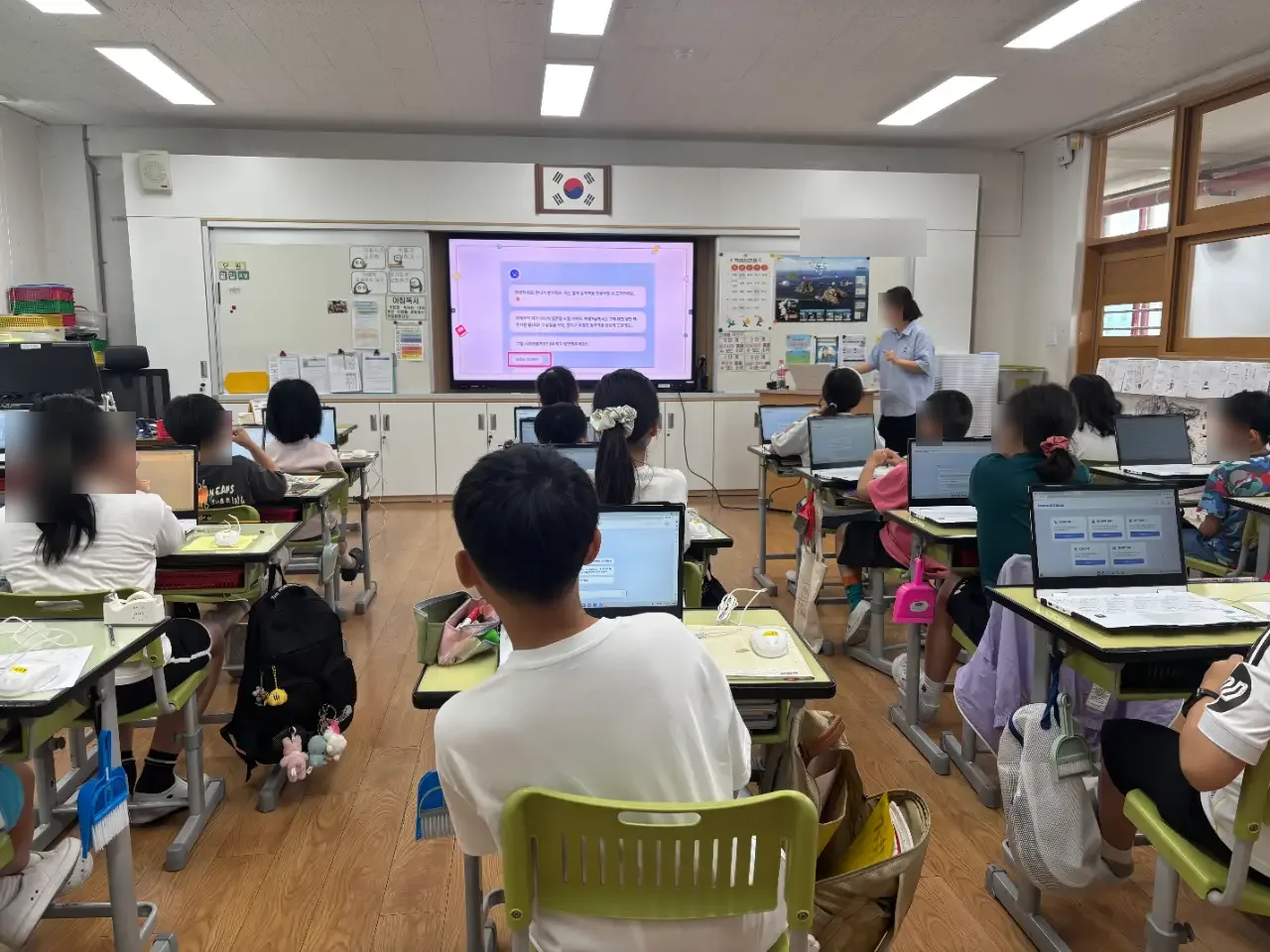 교실 앞 전자칠판과 강사, 자리에 앉은 학생들 (얼굴 흐림 처리)