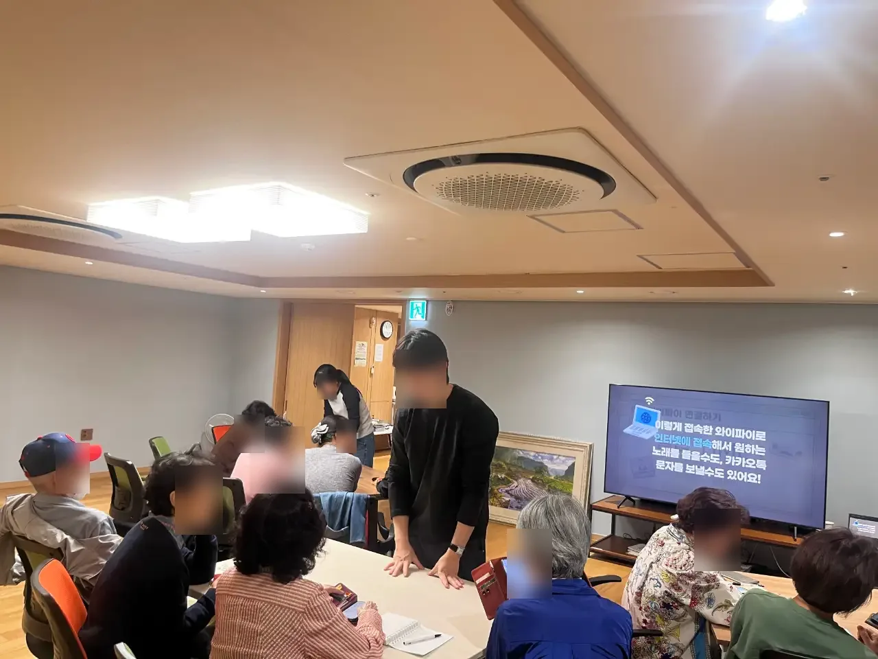 경로당에서 TV 화면을 보며 설명하는 봉사자와 어르신들 (얼굴 흐림 처리)