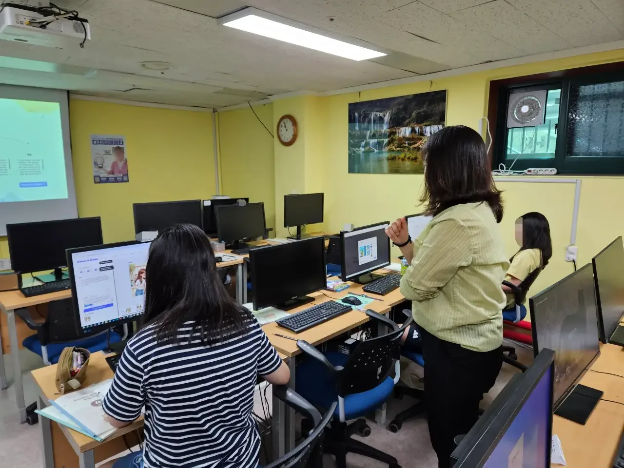 컴퓨터실에서 학생 옆에 서서 지도하는 강사 (얼굴 흐림 처리)